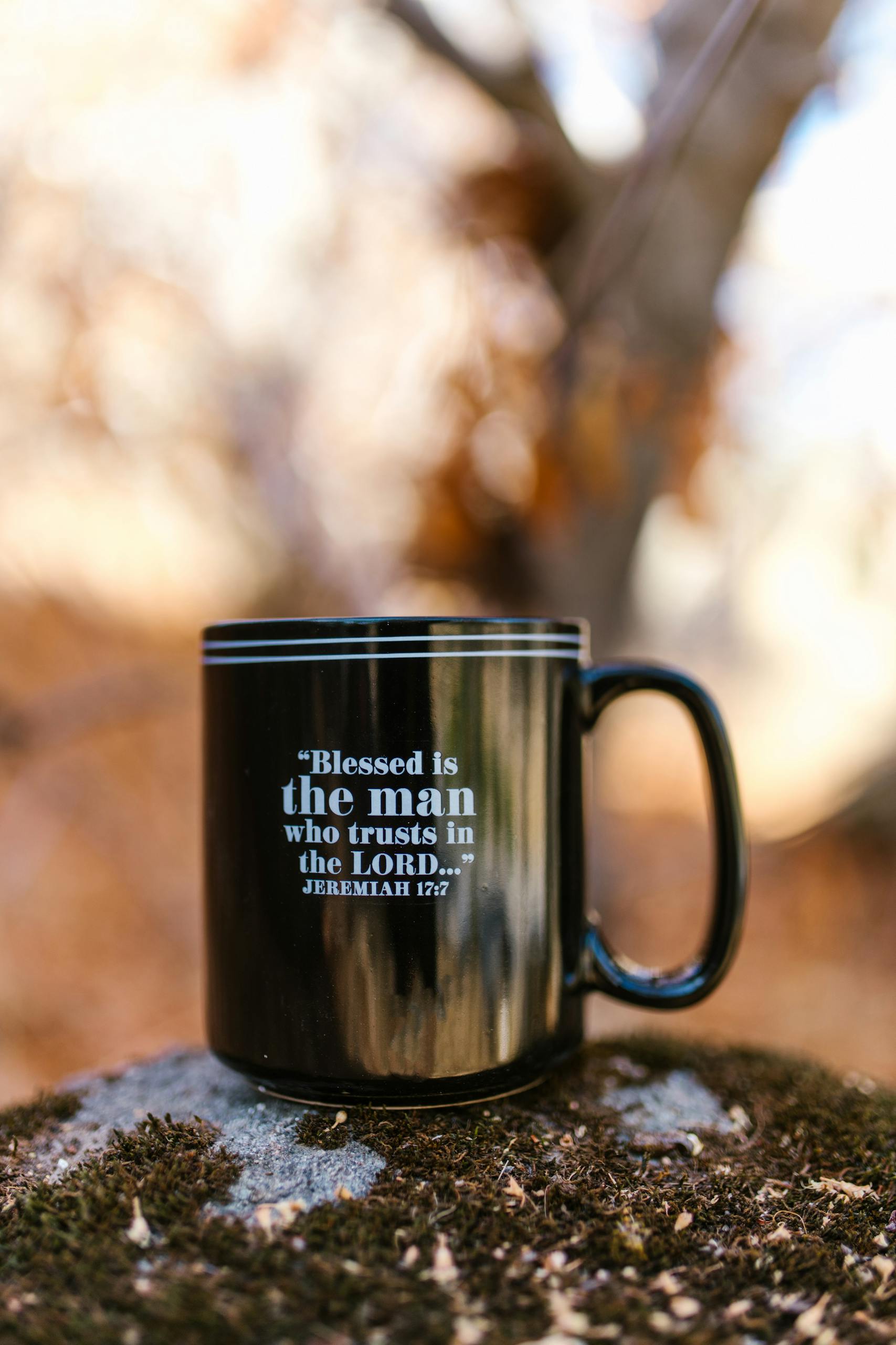 A black mug with a religious quote on a mossy rock, surrounded by autumn foliage.