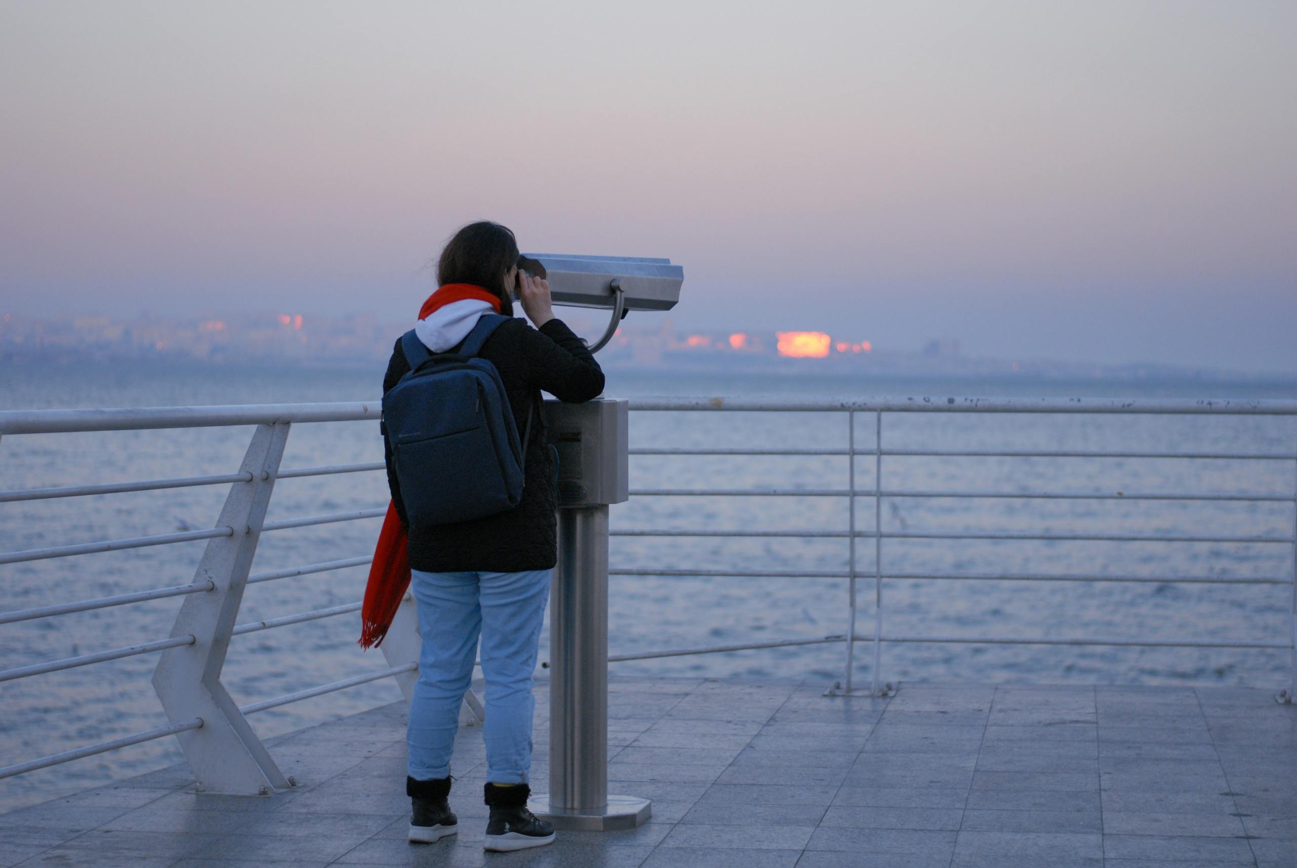 A woman with a backpack uses a tower viewer to observe the sea from a promenade at twilight.